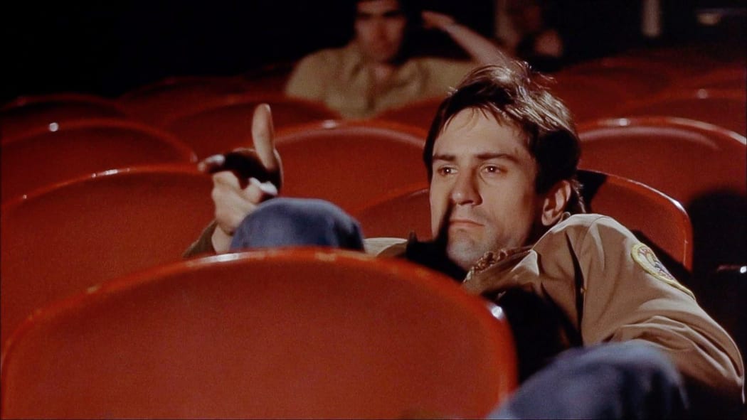 Movie still from Martin Scorsese's film Taxi Driver showing Robert De Niro as Travis Bickle slouching in a cinema with his fingers pointing like a gun barrel.